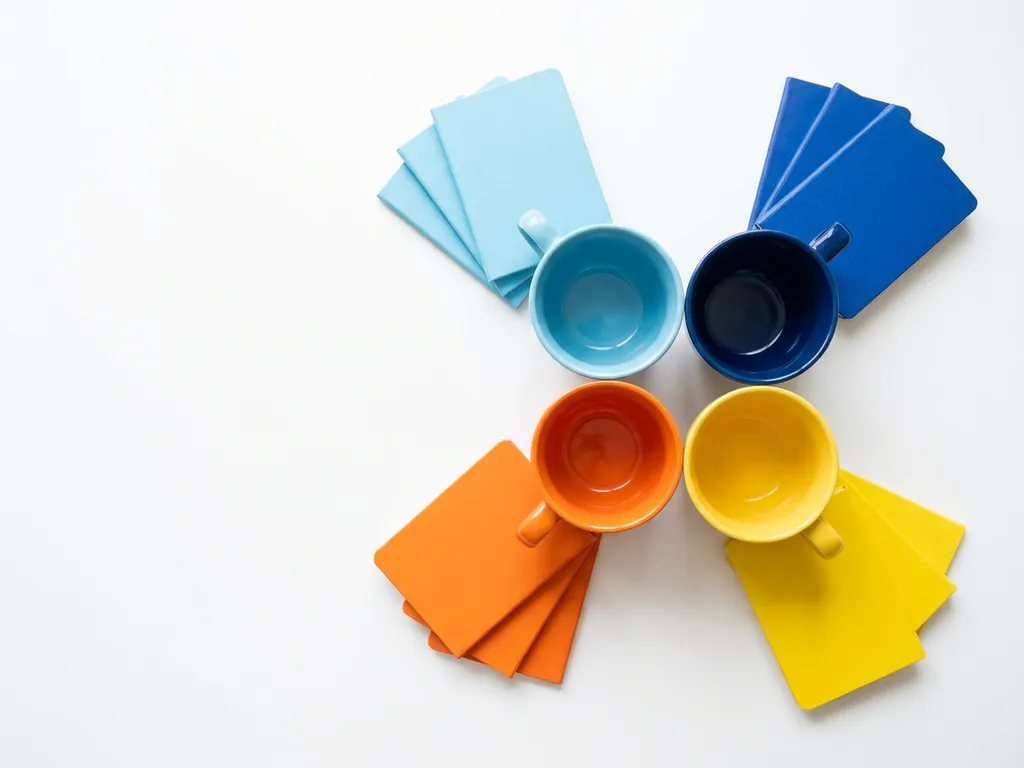 Kleurrijke koffiekopjes in lichtblauw, donkerblauw, oranje en geel gerangschikt met bijpassende notitieboekjes op wit bureau.